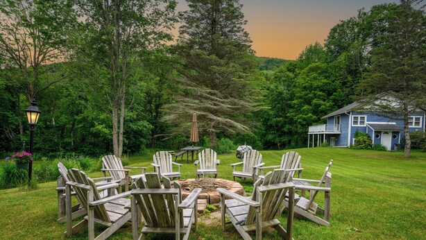 A circular arrangement of Adirondack chairs around a central fire pit in a lush, green outdoor setting.