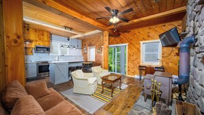 A cozy cabin interior with a wooden ceiling, a ceiling fan, a kitchen area, a living room with a sofa and armchairs, and a stone wall.