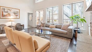 A spacious living room with a beige sofa, a glass coffee table, and a large window offering a view of the outdoors.