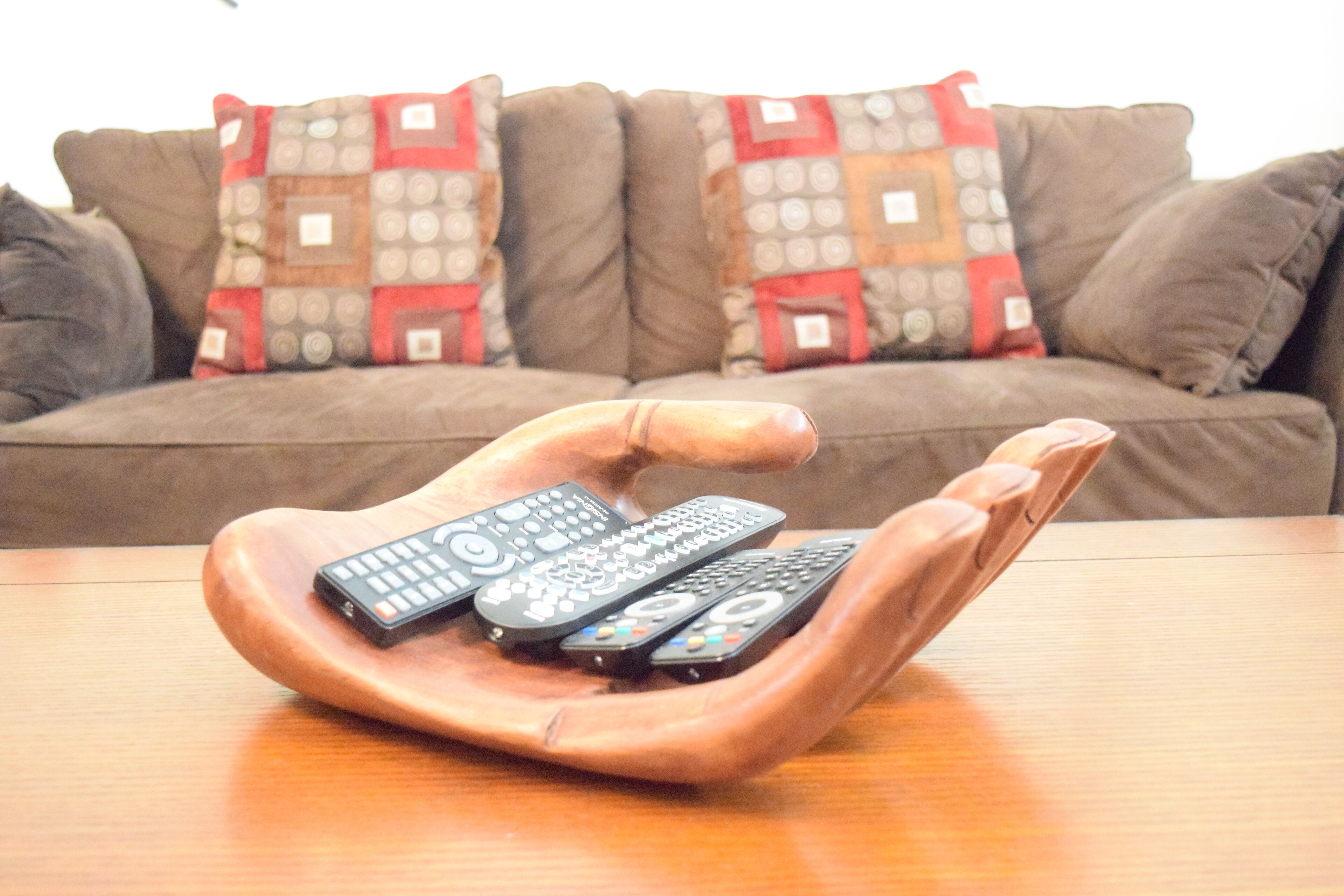 A wooden hand-shaped tray holding multiple remote controls on a wooden table.