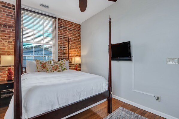 A bedroom with a four-poster bed, a brick wall, a TV mounted on the wall, and a window with blinds. - Heated Pool Access | 5*Clean | Flex Cancelation (Savannah)
