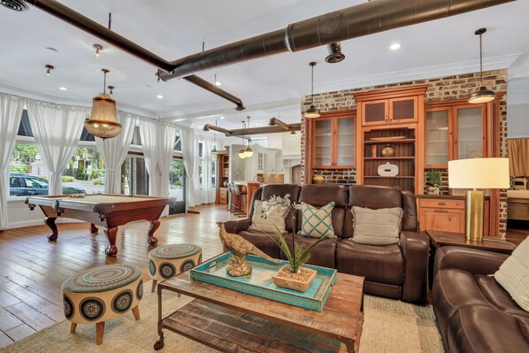 A spacious living room with a pool table, leather sofa, and a dining area with a brick wall and wooden cabinets. - Heated Pool Access | 5*Clean | Flex Cancelation (Savannah)