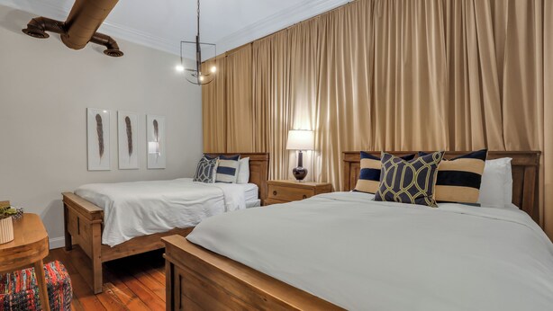 Une chambre d’hôtel avec deux lits simples, une tête de lit en bois, une table de chevet avec une lampe et un ventilateur de plafond.