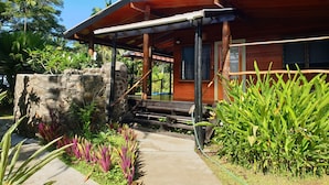 Une maison en bois avec un mur en pierre, une véranda et des plantes tropicales.