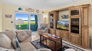A living room with a beige sofa, a wooden cabinet with glass doors, a television, and a view of a green landscape through a sliding glass door. - Remodeled! - Direct Walk Out to Beach & Pool - Pet Friendly  (Flagler Beach)