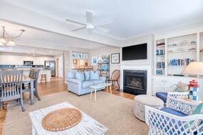 A spacious living room with a fireplace, a dining area, and a kitchen in the background. - Bacon in the Sun | 4BR with Ocean Views, Private Beach Access, Wraparound Porch in Quiet Area (Folly Beach)