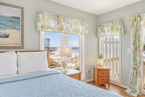 A bedroom with a bed, bedside table, lamp, and a view of the beach. - Bacon in the Sun | 4BR with Ocean Views, Private Beach Access, Wraparound Porch in Quiet Area (Folly Beach)