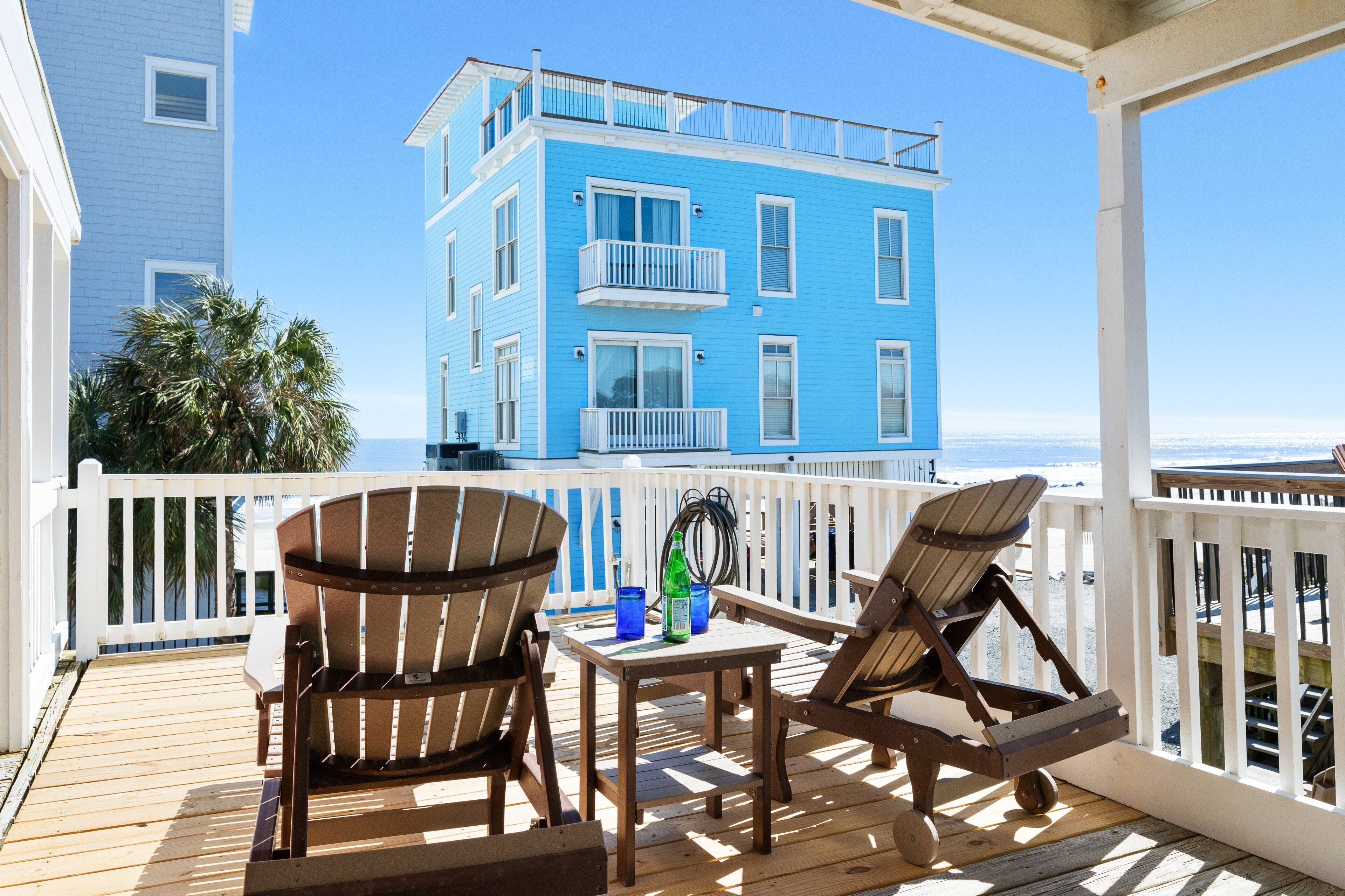 A wooden deck with two Adirondack chairs and a table, overlooking a blue building with balconies and a white fence.