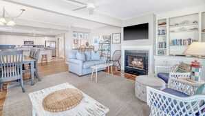 A spacious living room with a fireplace, a dining area with a kitchen in the background, and a bookshelf filled with books and decorative items.