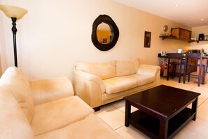 Un salon avec un canapé, une table basse, une lampe, un miroir et, au fond, un coin cuisine.