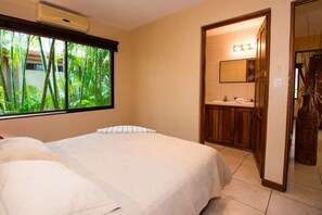 A hotel room with a bed, a window with a view of greenery, a bathroom with a mirror, and a sink. - Cozy Ground-Floor Condo w/ Community Pool, Rancho & Lush Garden Views (Santa Cruz)