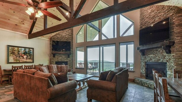 A spacious living room with a stone fireplace, large windows, and a high ceiling with exposed beams.