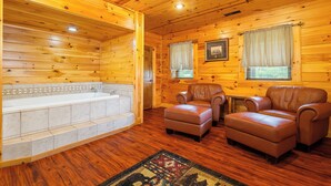 A log cabin interior with a wooden floor, a bathtub, two leather chairs, and a patterned rug.