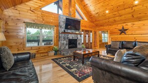 A log cabin interior with a stone fireplace, leather sofas, and a large TV.