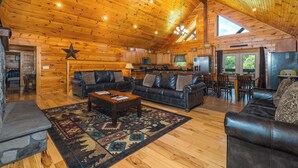 A spacious log cabin interior with a high ceiling, a stone fireplace, leather sofas, a dining area, and a kitchen.