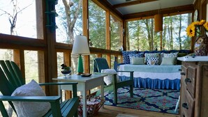 A cozy cabin with a wooden interior, a bed with patterned bedding, a table with a lamp, and a view of trees through large windows.