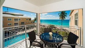 A balcony with a glass table, wicker chairs, and a view of the ocean and buildings. - Regal Beach Club #633 (Seven Mile Beach)