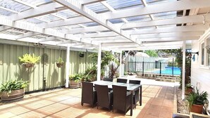 Une terrasse couverte avec une table et des chaises, des plantes en pot et une vue sur une piscine.