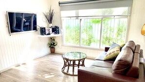 Un salon avec un canapé en cuir marron, une table basse en verre, un téléviseur à écran plat fixé au mur et une grande fenêtre donnant sur un paysage verdoyant.