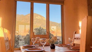 Un comedor con una mesa de madera dispuesta para cuatro, una araña y un gran ventanal con vistas a las colinas.