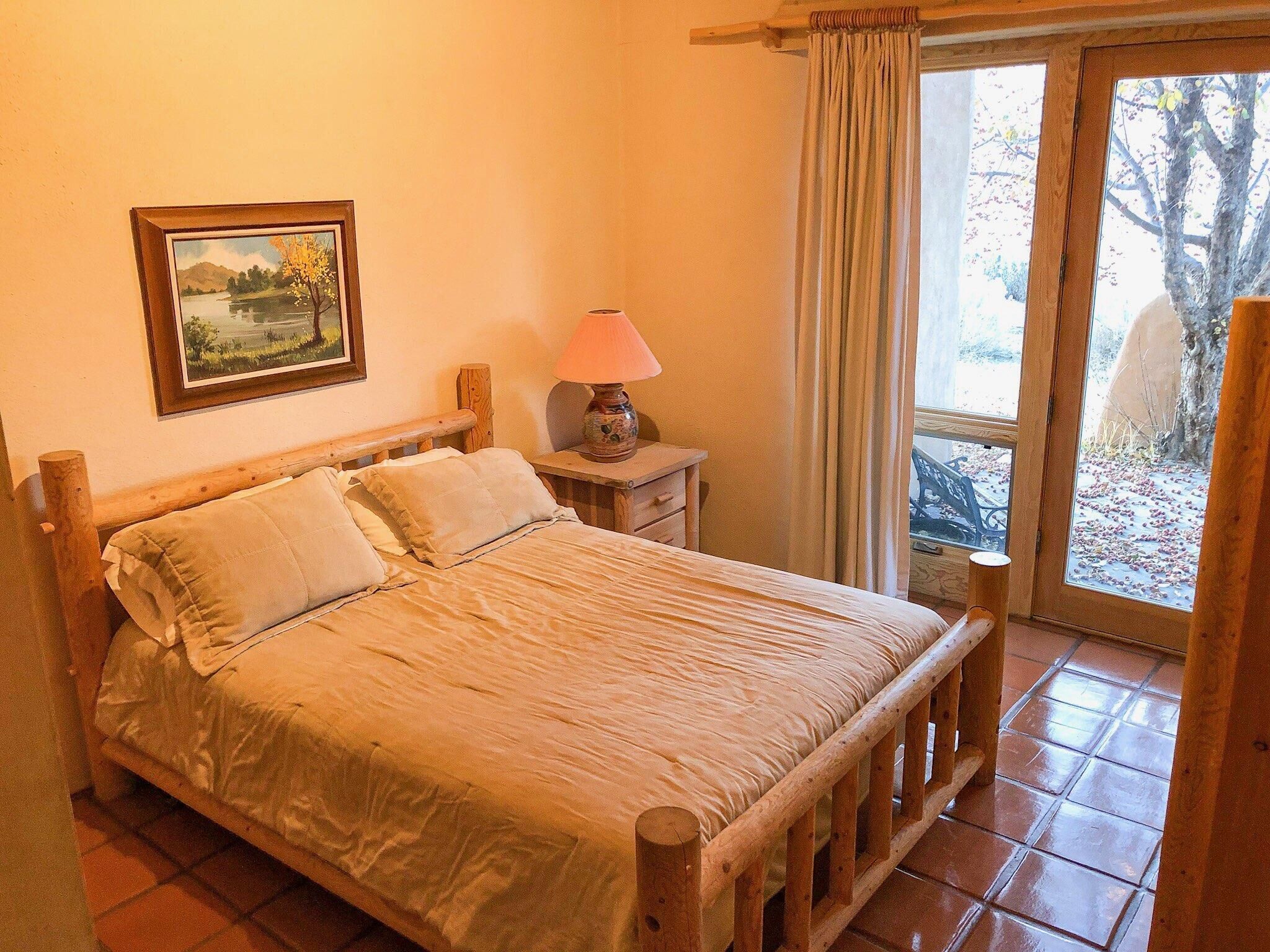 A bedroom with a wooden bed, a nightstand with a lamp, a window with curtains, and a framed picture on the wall.