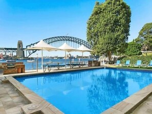 A swimming pool with lounge chairs and umbrellas, a large tree, and a view of a bridge in the background. - Harbourside7, Luxury 1 Bed Apartment (McMahons Point)