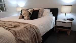 Ein ordentlich bezogenes Bett mit einem gemusterten Überwurf, ein Nachttisch mit einer Lampe und eine digitale Uhr, die die Uhrzeit anzeigt.