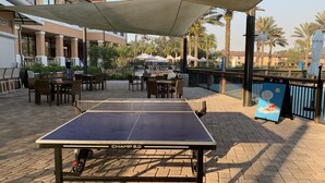 Un tavolo da ping pong con l'etichetta 'CHAMP 5.0' sotto un'area ombreggiata, con posti a sedere all'aperto e una piscina sullo sfondo.