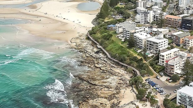 Vista aérea de uma área costeira com praia, linha de costa rochosa e edifícios residenciais.