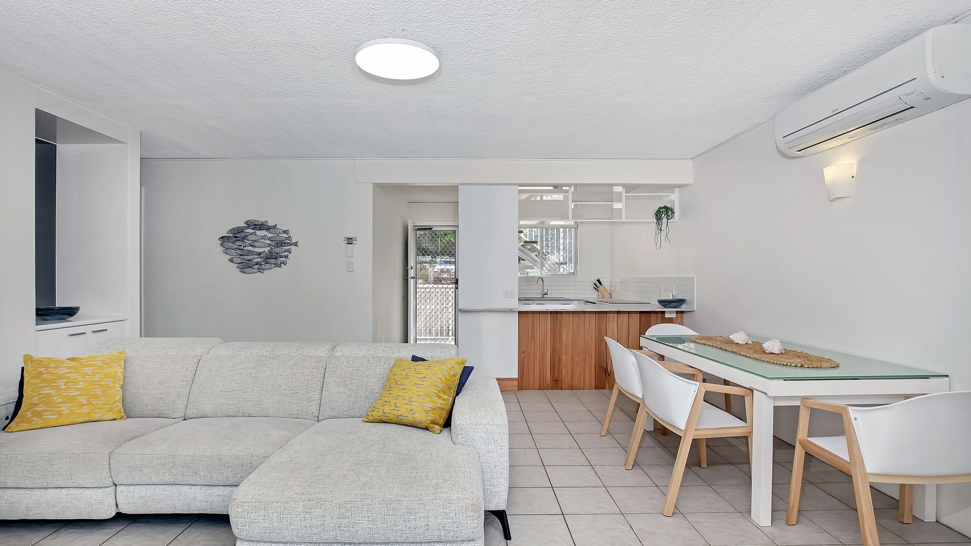 Ein modernes Wohnzimmer mit einer Couch und Sesseln, ein Essbereich mit einem Glastisch und eine durch eine offene Tür sichtbare Küche.