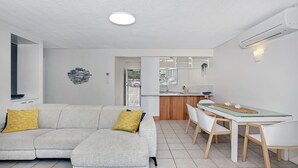 Uma sala moderna com sofá e poltronas, uma área de jantar com mesa de vidro, e uma cozinha visível através de uma porta aberta.