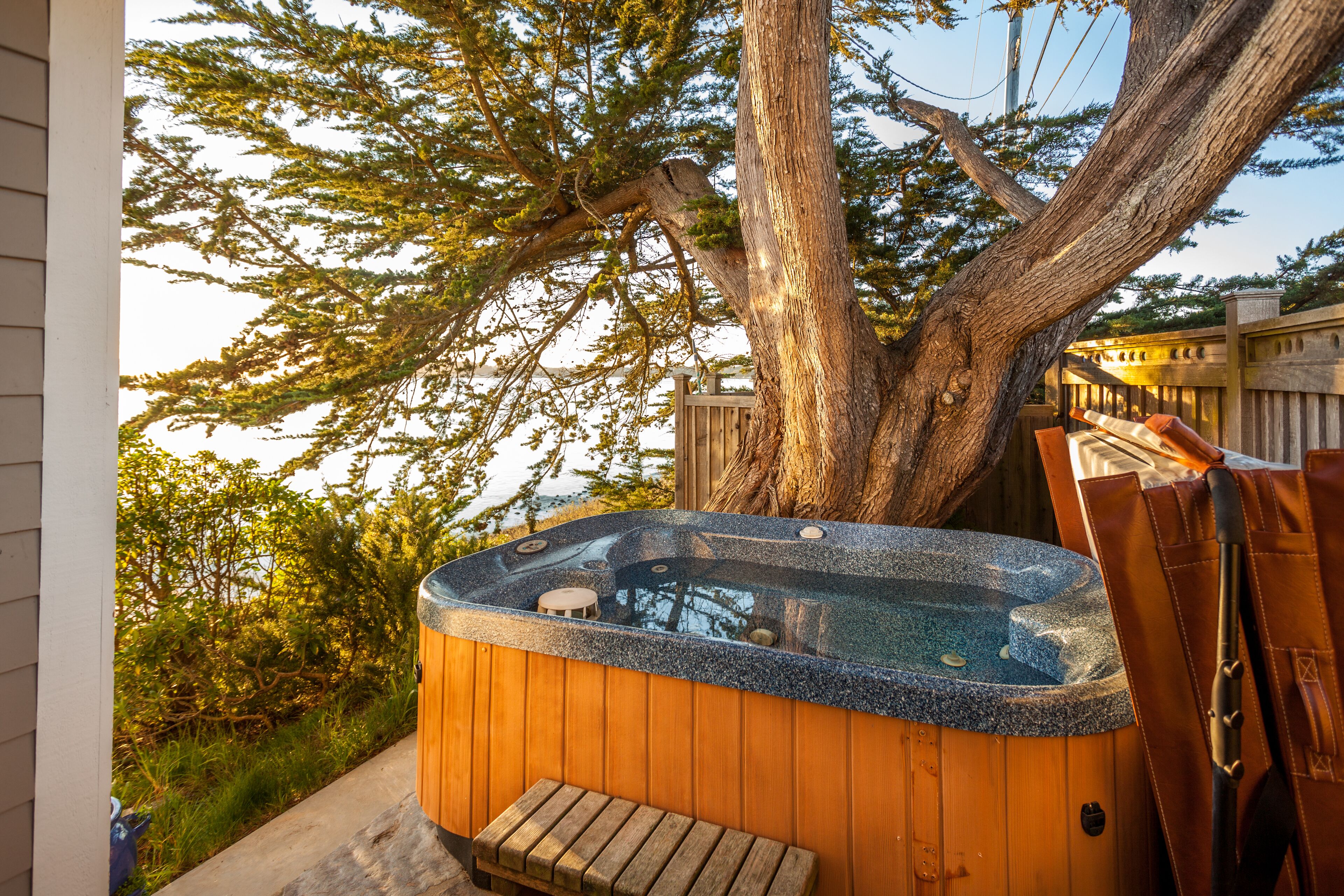A hot tub with a wooden deck, a large tree, and a wooden fence.