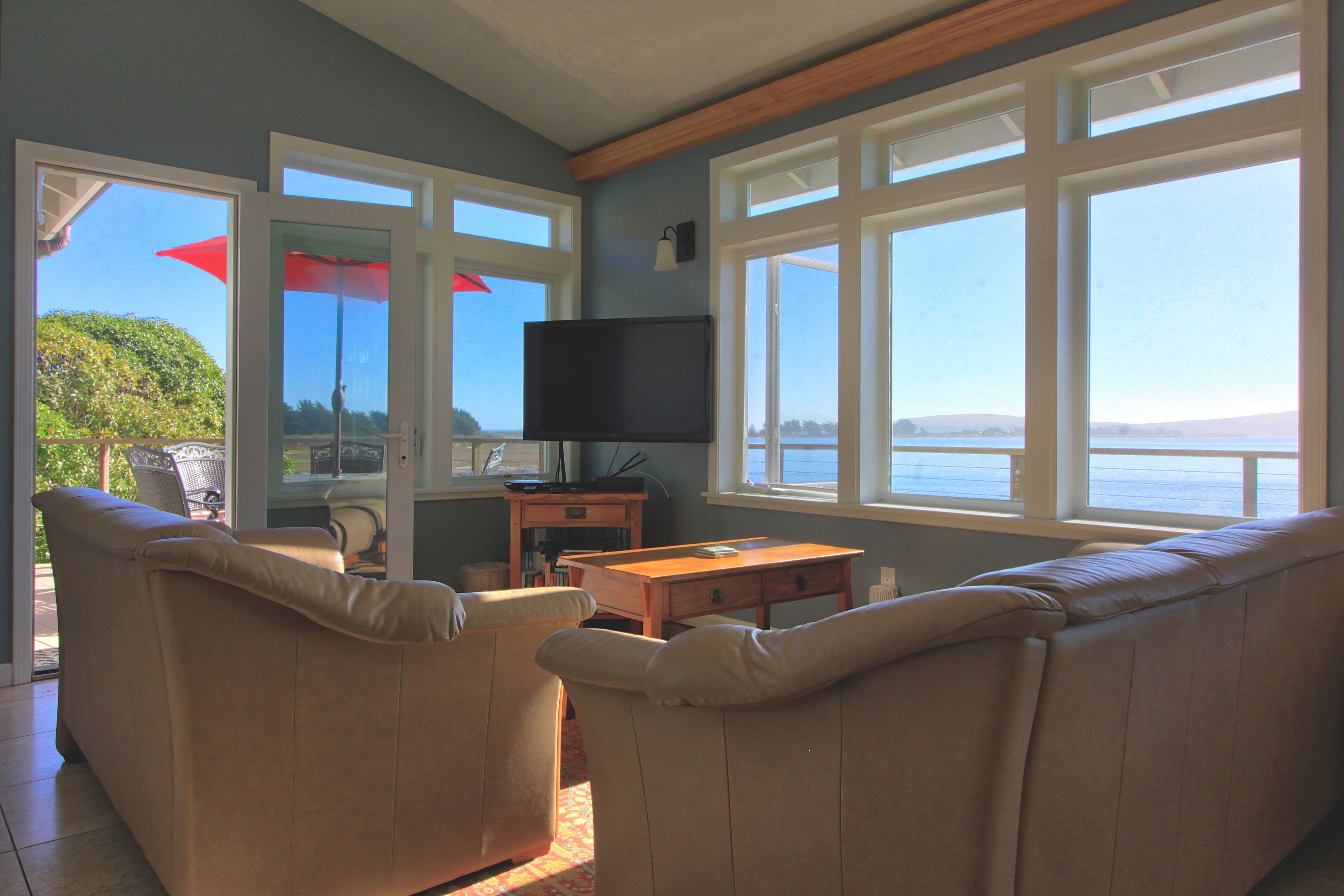 A living room with a TV, beige sofas, a wooden table, and large windows overlooking a body of water.