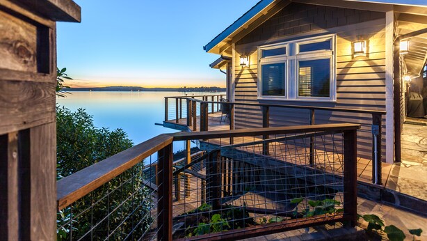 A wooden cabin with a deck overlooking a calm lake, featuring a staircase and a railing. - Bird Watch @ Land's End with Amazing Views of the Harbor and Pacific Ocean (Bodega Bay)