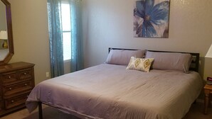 Een slaapkamer met een bed, een dressoir, een nachtkastje, een lamp, een schilderij en een raam met gordijnen.
