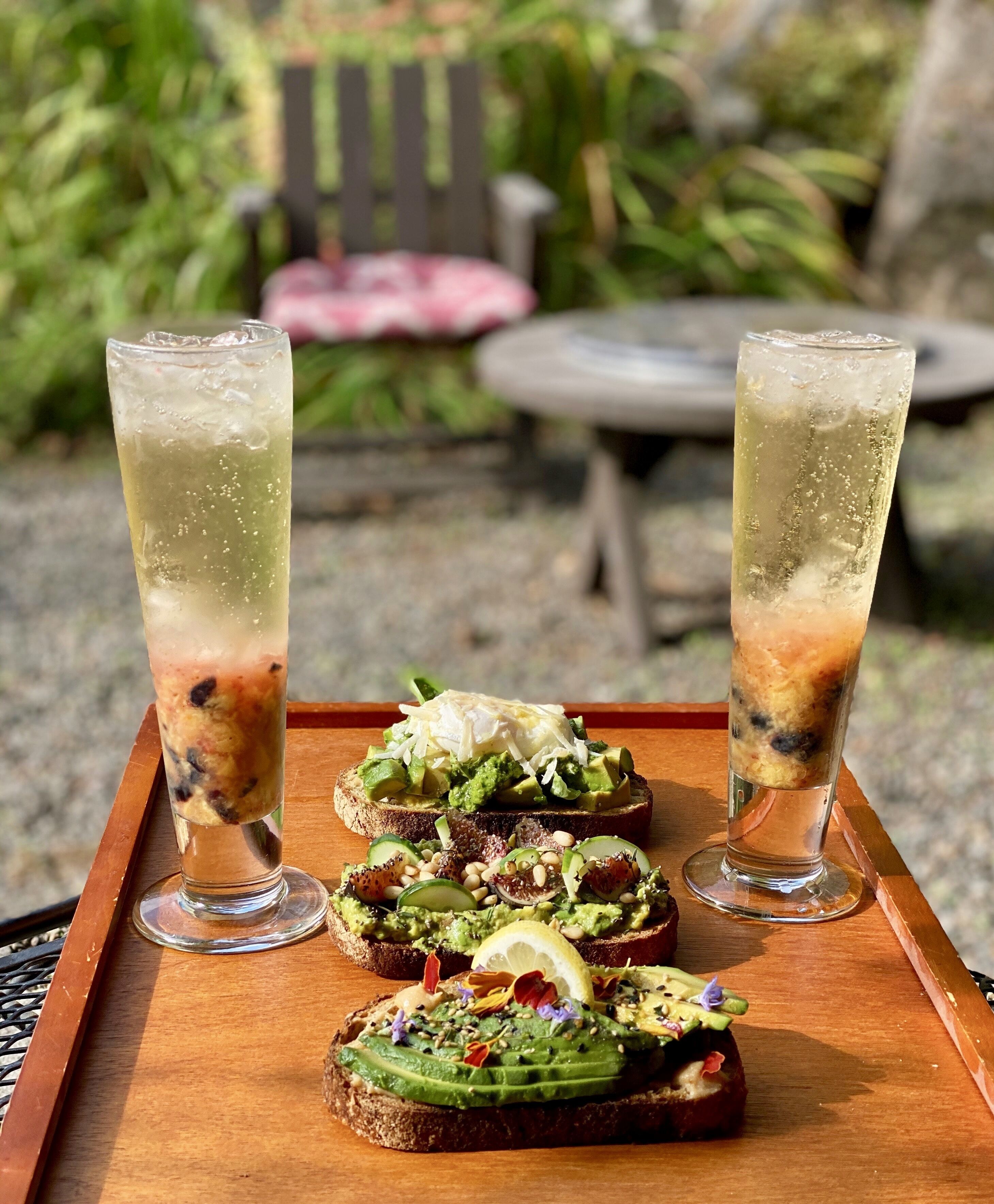 Deux grands verres de boisson glacée, avec une boisson à plusieurs couches, deux tranches de pain grillé garnies d’avocat et de divers accompagnements, et un plateau en bois.