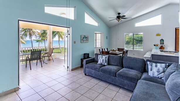 A spacious living area with a large gray sofa, a dining table, and a view of the ocean through a sliding glass door.