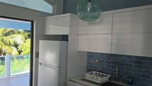 A kitchen with white cabinets, a blue backsplash, a hanging light fixture, and a view of palm trees through a door.