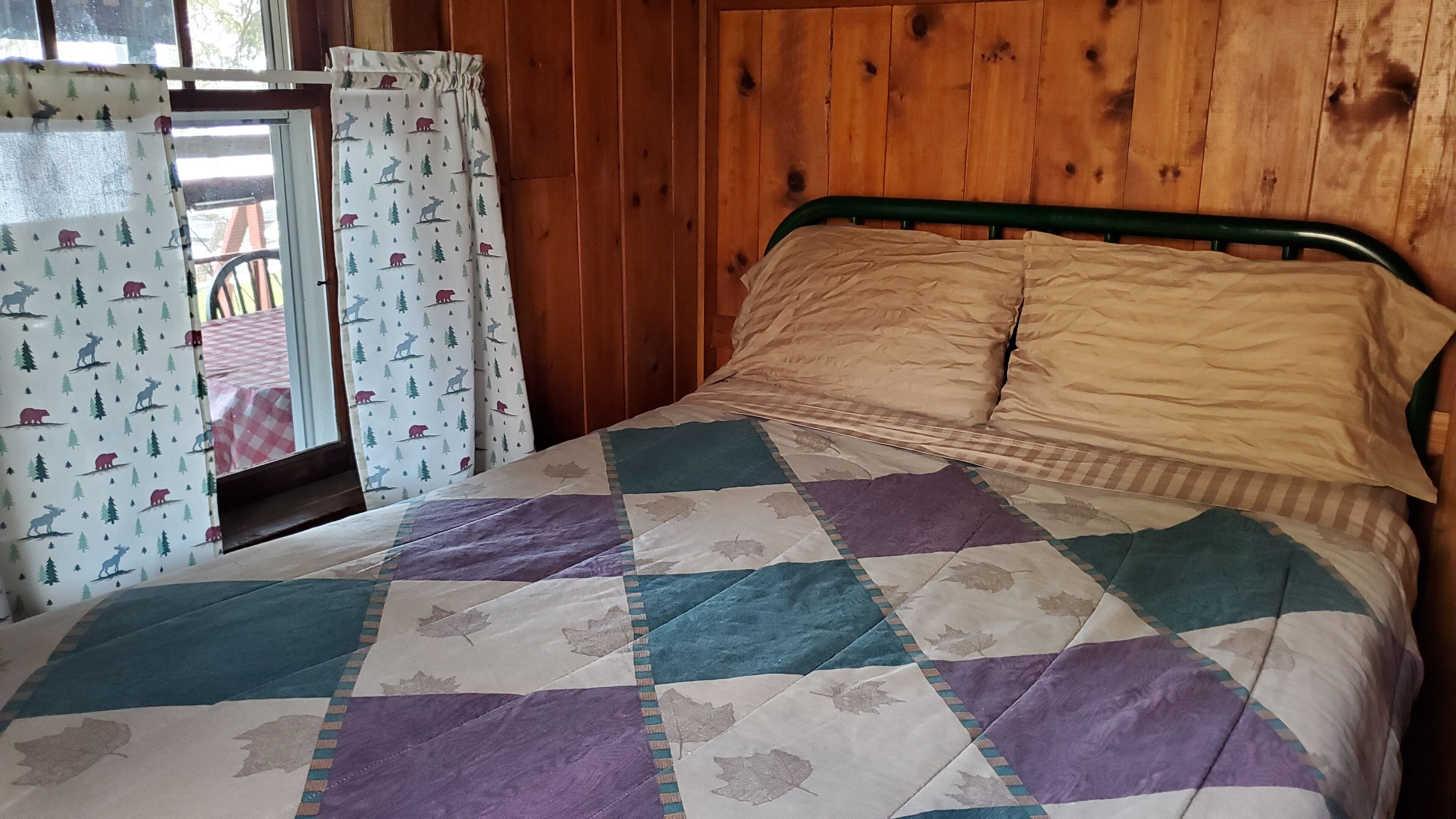 A bed with a quilt, wooden walls, and a window with curtains.