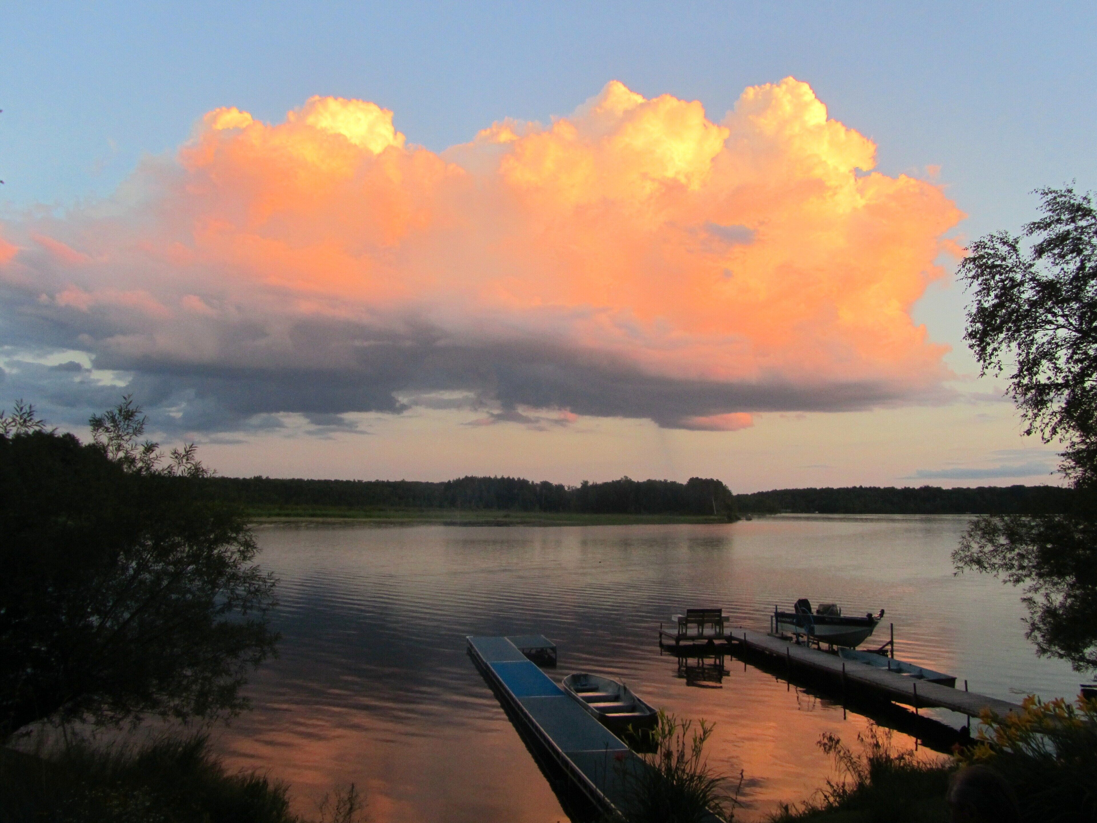 A calm lake with a dock and boats, surrounded by trees and a cloudy sky.