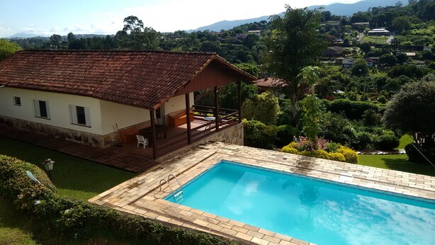 A pool with a tiled deck, a small building with a tiled roof, and a view of a lush landscape with trees and hills in the background. - House with 5 bedrooms, beautiful view of Pedra Grande. (Atibaia)