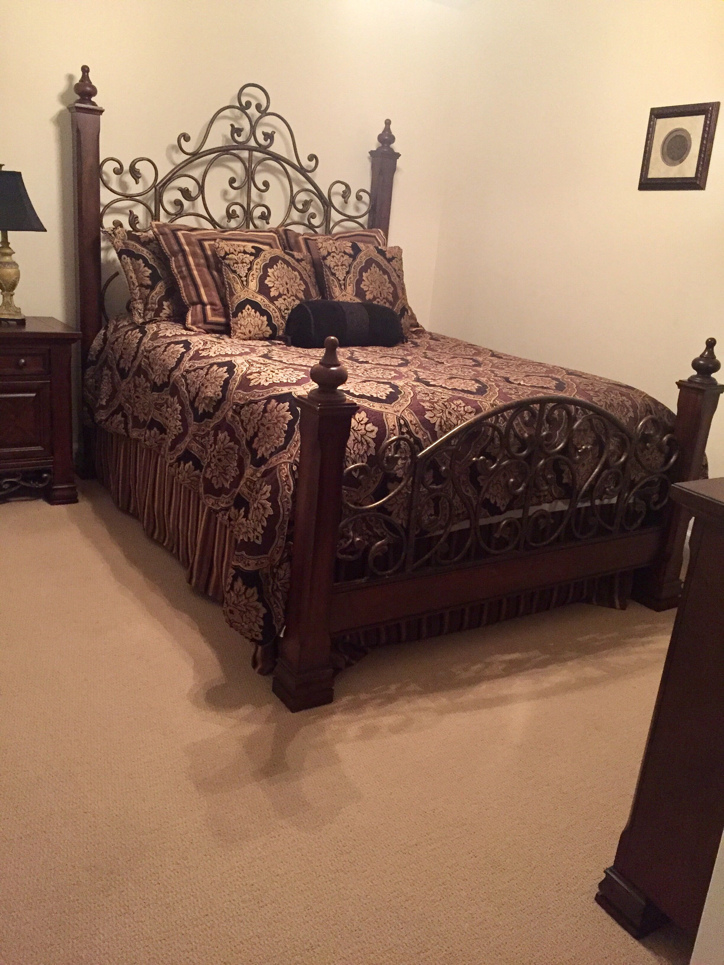 A bed with an ornate metal headboard, a dark patterned bedspread, and a wooden nightstand with a lamp.