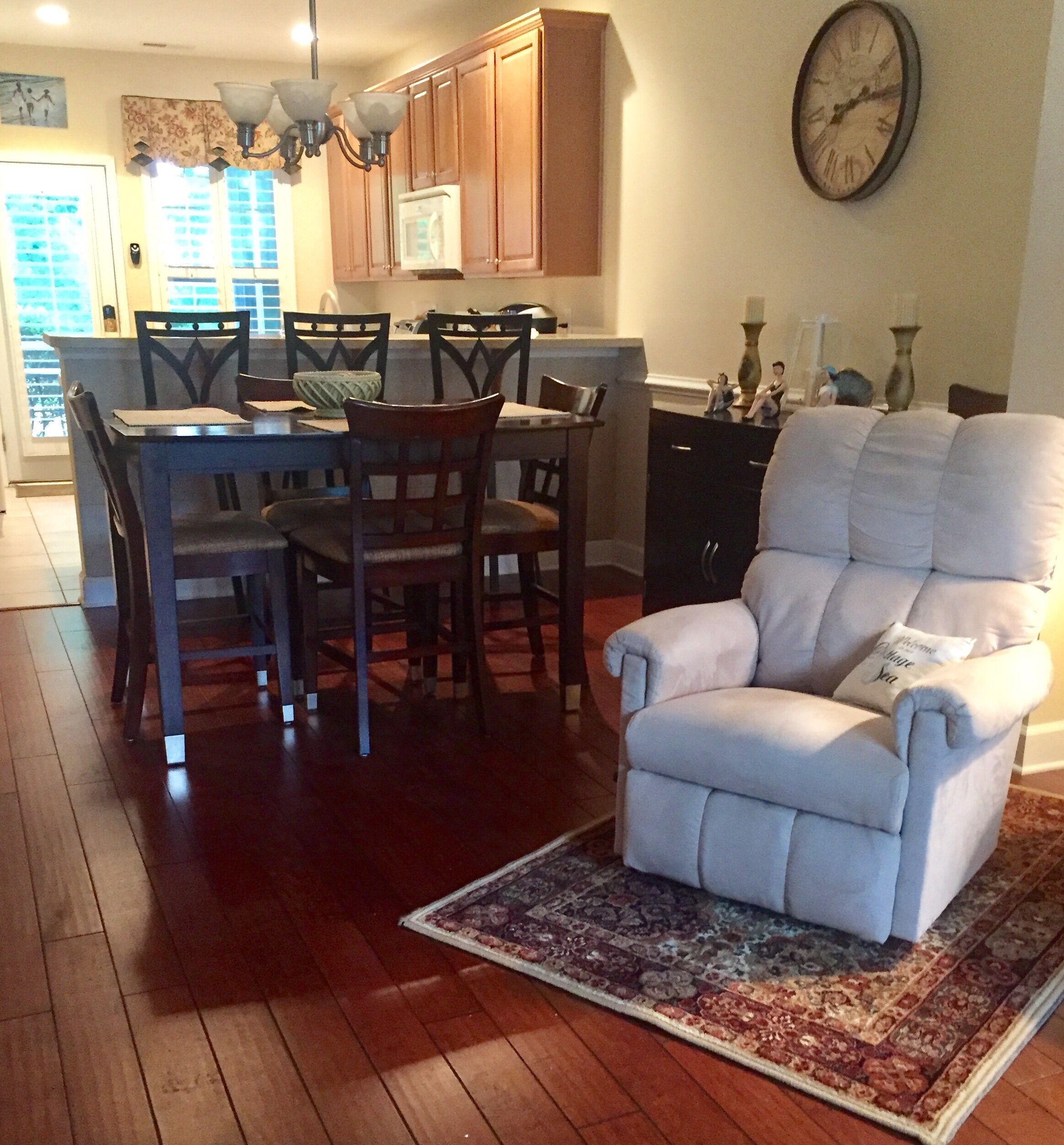 A dining area with wooden chairs and a table, a white armchair with a throw pillow, and a patterned rug on a wooden floor.