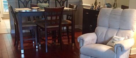 A dining area with wooden chairs and a table, a white armchair with a throw pillow, and a patterned rug on a wooden floor.