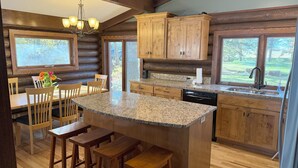 A log cabin interior with a kitchen featuring wooden cabinets, granite countertops, and a dining area with wooden chairs and a table. - Remodeled Log Cabin Home Chain of Lakes (Alexandria)
