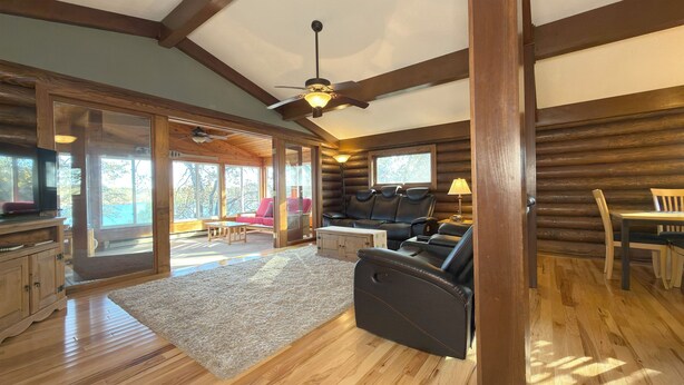 A log cabin interior with a wooden ceiling, a ceiling fan, a fireplace, leather seating, and large windows. - Remodeled Log Cabin Home Chain of Lakes (Alexandria)