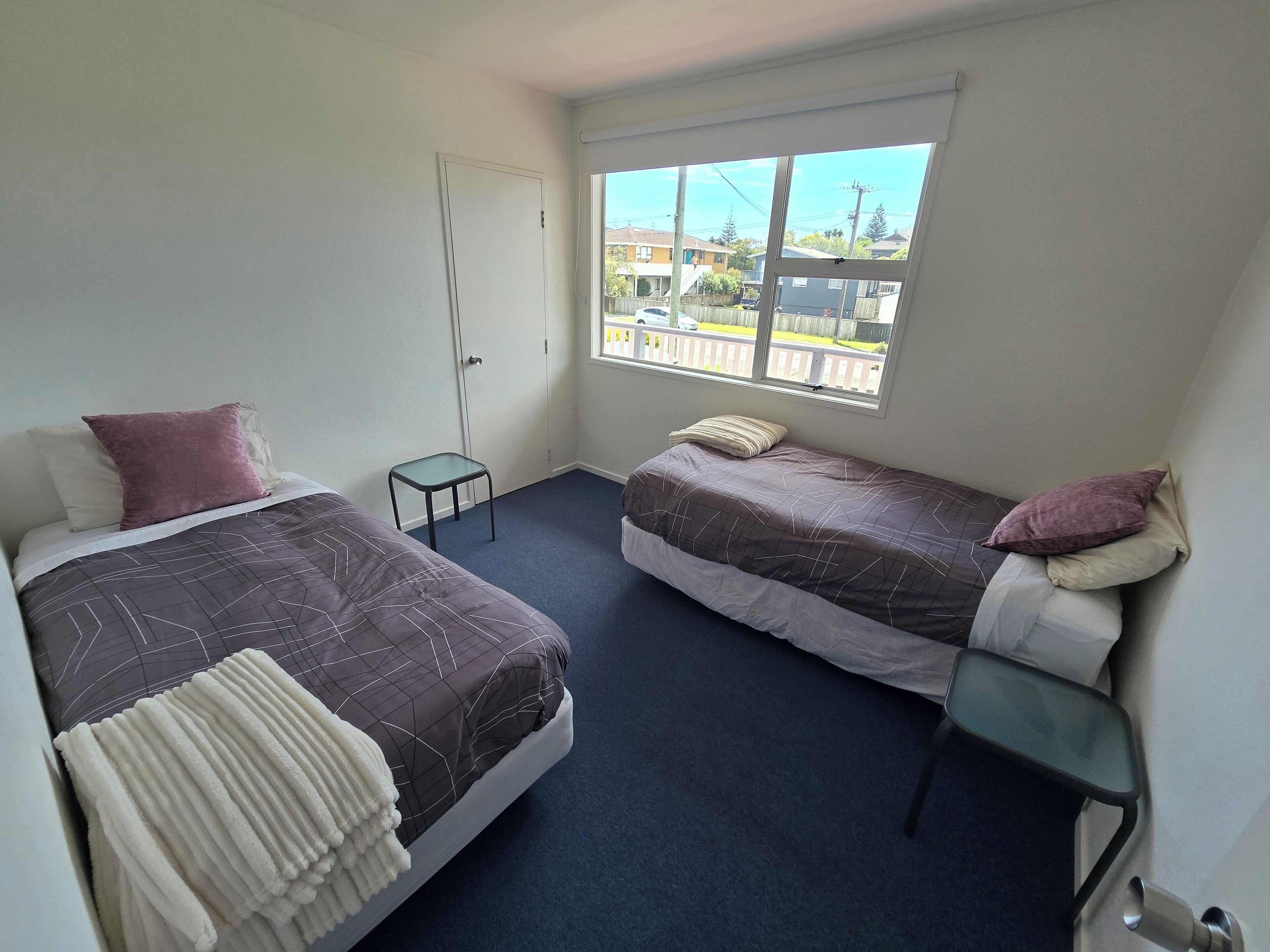 Ett litet, rent rum med två sängar, ett litet bord och ett fönster med utsikt över ett bostadsområde.