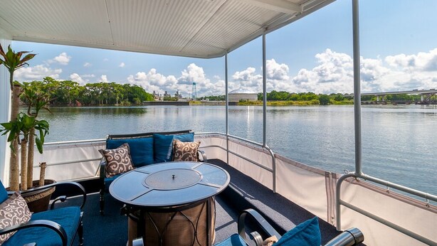 A covered outdoor seating area with a table and chairs, overlooking a calm body of water with a bridge in the background.