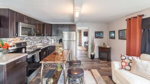 A modern kitchen with dark wood cabinets, stainless steel appliances, and a marble countertop. There is a dining area with a table and stools, and a living room with a sofa and a fireplace.