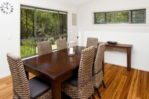 Comedor con mesa de madera y sillas tejidas, ventanas amplias con vistas a zonas verdes y un reloj de pared.